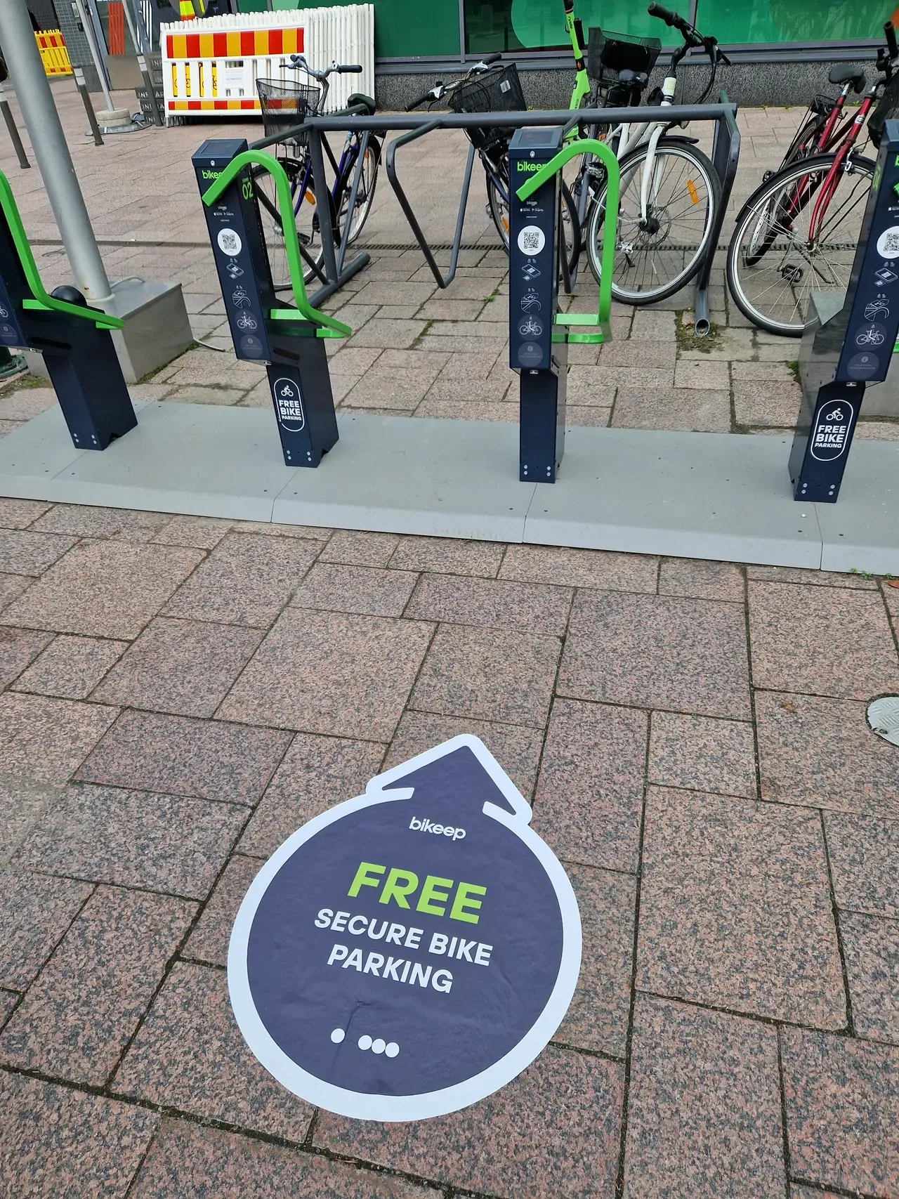 A Bikeep floor decal reading "Free secure bike parking" beside a station with bikes docked in it, at a shopping centre in Finland